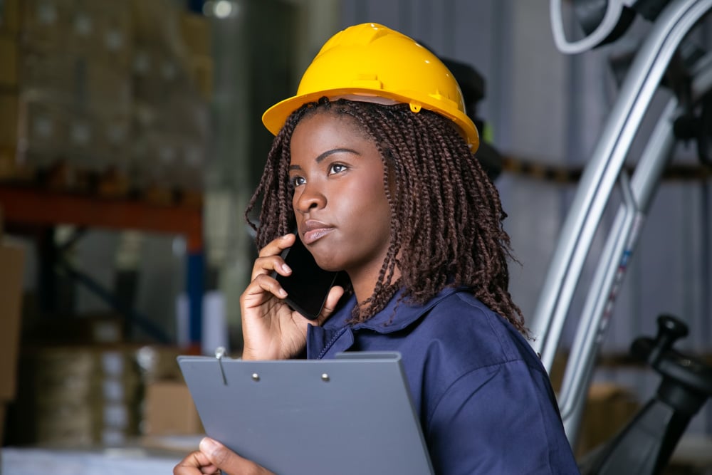 pensive-black-female-engineer-hardhat-standing-warehouse-talking-cellphone-shelves-with-goods-background-copy-space-labor-communication-concept